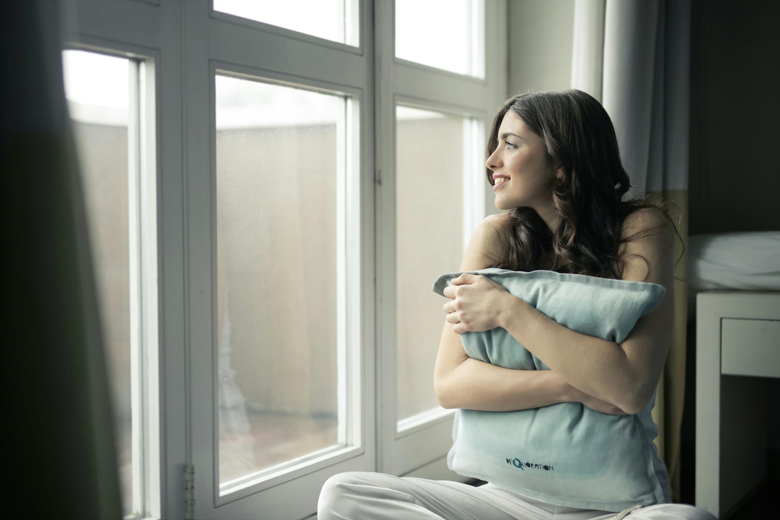 smiling woman hugging a blue pillow, looking out a window