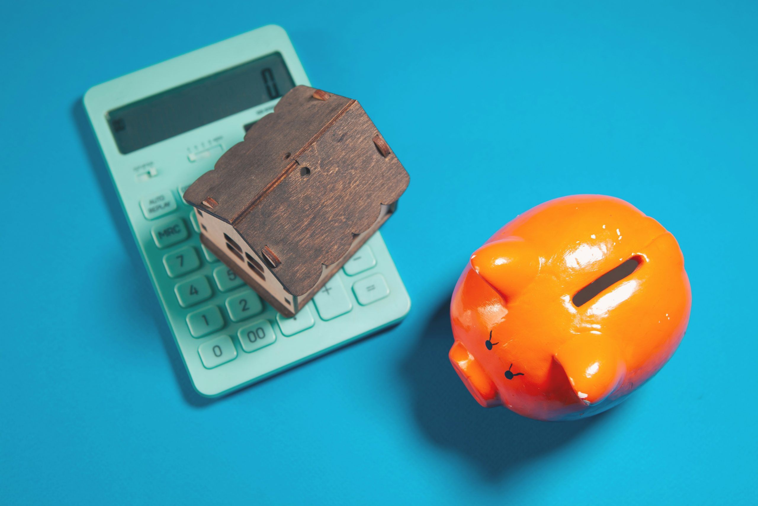 miniature house resting on a calculator, next to an orange piggy bank